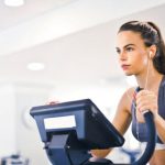Cardio - Serious fit woman in earphones and activewear listening to music and running on treadmill in light contemporary sports center