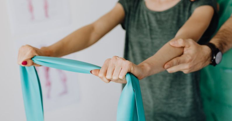 Rehabilitation - Crop unrecognizable orthopedist in uniform and wristwatch supporting shoulder and reached arm of faceless female patient with resistance band in doctor office